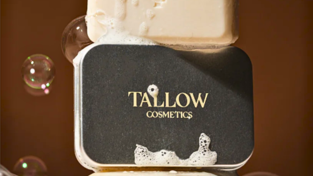 Tallow Cosmetics soap tin with a foaming soap bar on top, floating bubbles, on a warm brown background.